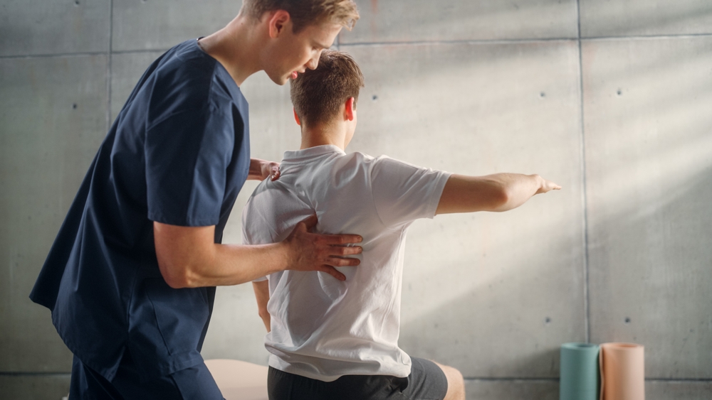 A physical therapist touching a man's back who is kneeling on the floor.
